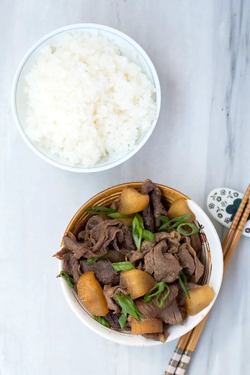 Top view of a bowl of beef with daikon stir fry with a bowl of rice and chopsticks next to it. 