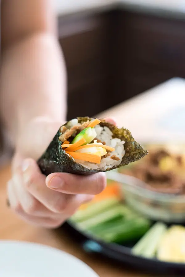 Holding a canned tuna sushi hand roll with ingredients in background.