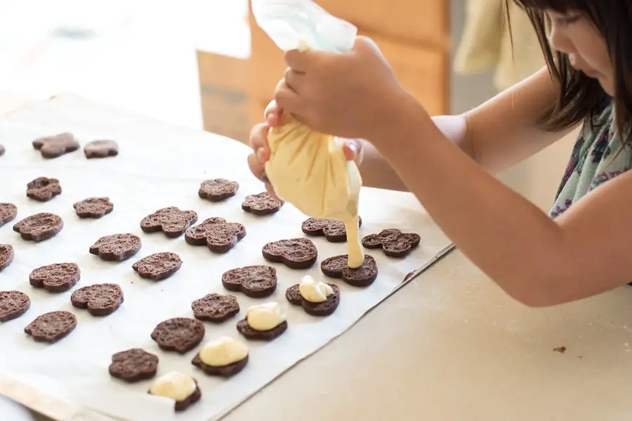 Child piping orange buttercream onto baked chocolate sandwich cookies.