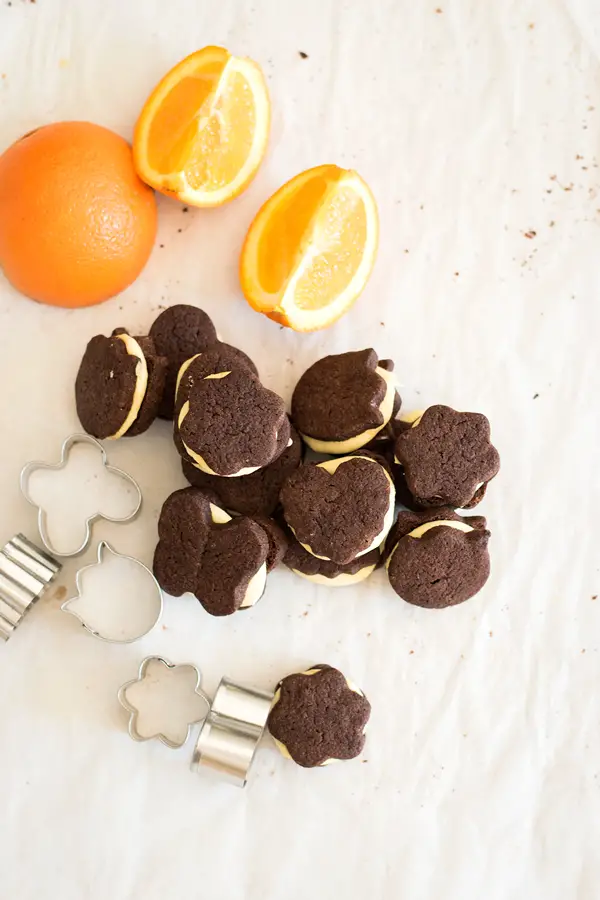 Top view of chocolate orange sandwich cookies with sliced oranges and mini cookie cutters.