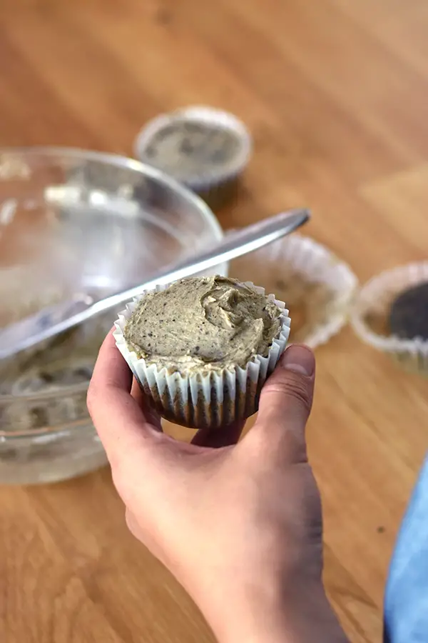 Holding a black sesame cupcake, with bowl of peanut butter frosting in the background. 