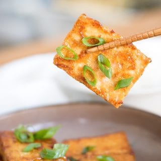 chopsticks holding soy sauce and butter glazed tofu with green onion garnish.