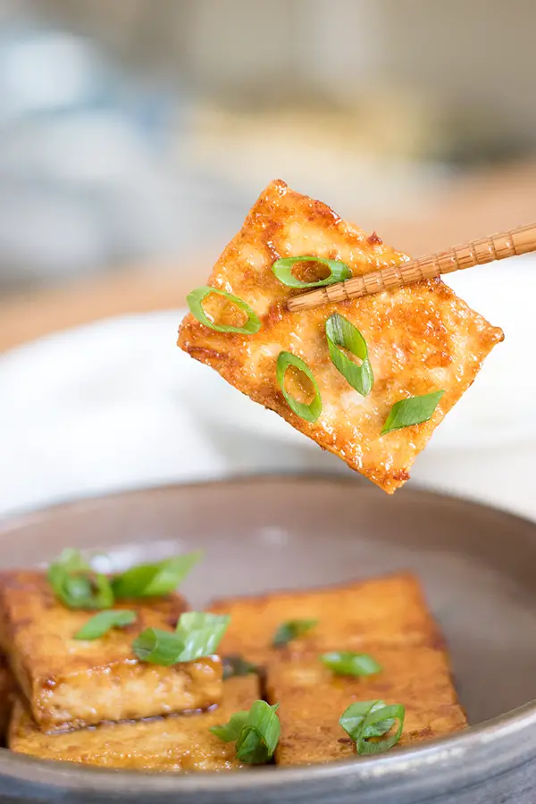 chopsticks holding soy sauce and butter glazed tofu with green onion garnish.