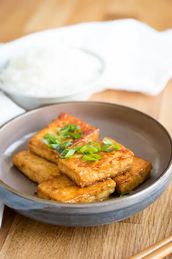 Plate of pan fried tofu, garnished with sliced green onion, and with a bowl of rice.